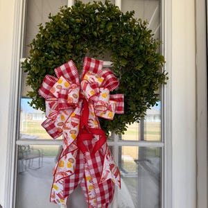May include: A green wreath with a red and white checkered bow and red ribbons. The bow features a pattern of crawfish, lemons, and other food items. The wreath is hanging on a white door with a window.