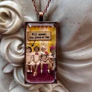 May include: A rectangular pendant necklace featuring a copper-toned metal frame and a chain. The pendant displays a vintage-style photograph of four children sitting in a row, with a yellow banner above them containing the text Will answer the phone at 2am.