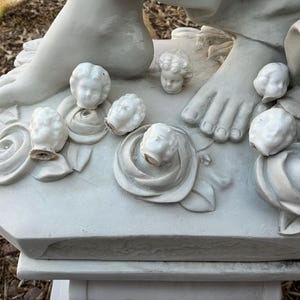 May include: A close-up of a white marble statue featuring a foot, cherub heads, and rose details. The statue is weathered, with visible details of the cherub faces and rose petals. The statue is on a white base.