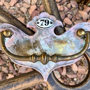 May include: A distressed, ornate drawer pull with a brass handle and a decorative backplate. The backplate is painted in shades of pink, blue, and gold, and features the number "79" on a small, oval plaque.