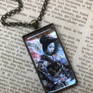 May include: A vintage-style necklace featuring a rectangular glass pendant with a colourful image of a woman in a traditional Japanese kimono. The woman is holding a bouquet of flowers and has a serene expression on her face. The necklace is made of a brass chain.