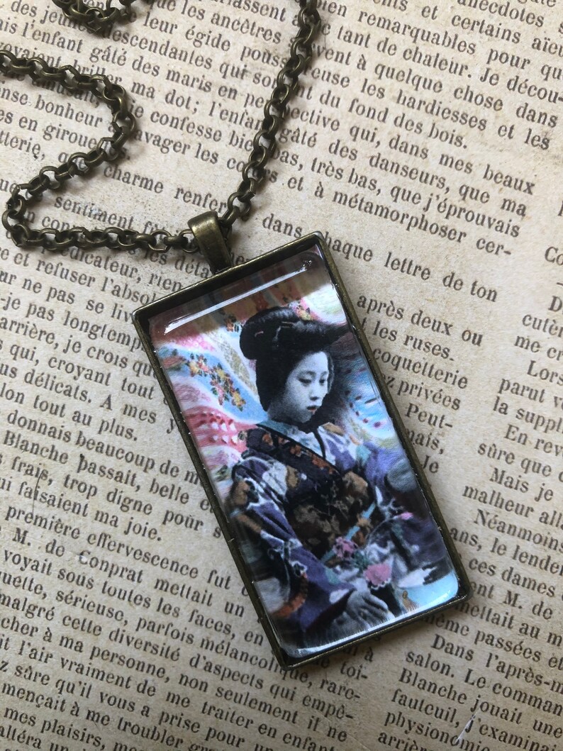 May include: A vintage-style necklace featuring a rectangular glass pendant with a colourful image of a woman in a traditional Japanese kimono. The woman is holding a bouquet of flowers and has a serene expression on her face. The necklace is made of a brass chain.