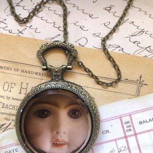 May include: A gold-toned necklace with a round pendant featuring a vintage-style magnifying glass. The glass reveals a doll's face with brown eyes and a slightly open mouth. The necklace is on a piece of paper with handwritten text.