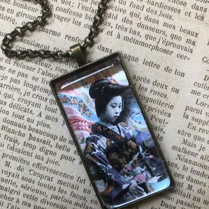 May include: A vintage-style necklace featuring a square glass pendant with a colourful image of a woman in a traditional Japanese kimono. The woman is holding a bouquet of flowers and has a serene expression on her face.