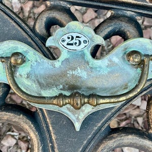May include: Antique bronze drawer pull with a decorative backplate and a curved handle. The backplate has a turquoise patina and a small, oval tag with the number 25. The handle is a warm bronze color.