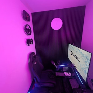 May include: A gaming setup with a racing simulator theme. The room features a racing seat, steering wheels, and a monitor displaying "WRC" graphics. The walls are accented with a black panel and a circular light.