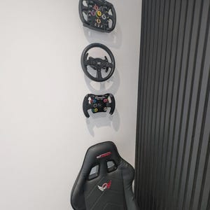 Può includere: Una sedia da gaming nera con dettagli rossi e un logo, sotto tre volanti da corsa montati. I volanti hanno vari design, con pulsanti e comandi. Il muro è bianco con un pannello nero.