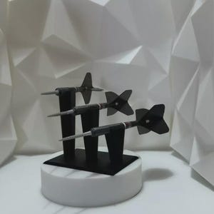 May include: Three darts with black flights and silver barrels are displayed on a black stand atop a white pedestal. The darts are set against a white, geometric patterned backdrop, creating a clean, modern aesthetic.