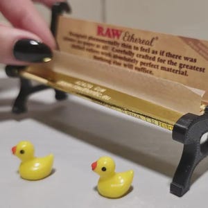 May include: A miniature black bench with a gold seat holds a package labelled "RAW Ethereal". Two small yellow duck figurines with red beaks sit in front of the bench. The text on the package reads "RAW Ethereal".