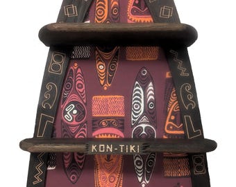 Puede incluir: Una estantería de pared de madera con forma de tabla de surf y tres niveles horizontales. El marco de madera oscura presenta patrones geométricos grabados y un motivo solar. El panel trasero muestra un patrón repetitivo de máscaras tiki estilizadas en naranja, rosa y blanco sobre un fondo granate. El estante central incluye una placa con el texto KON-TIKI.
