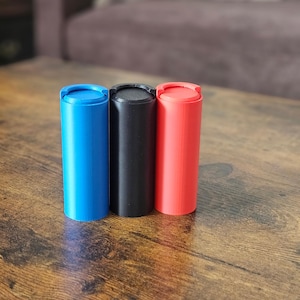 May include: Three cylindrical containers with lids, one blue, one black, and one red, sitting on a wooden surface.