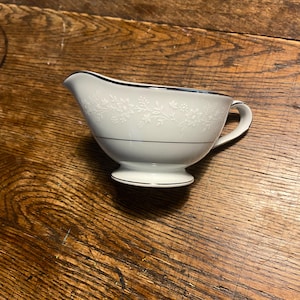May include: A white porcelain gravy boat with a floral pattern and a silver trim. The gravy boat has a spout, a handle, and a pedestal base. The design is a classic style, suitable for formal dining.