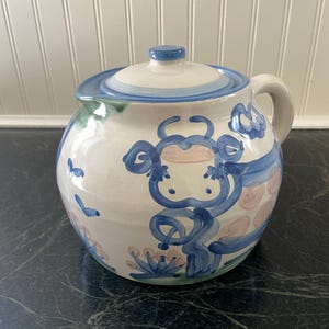 May include: A ceramic, lidded jar with a whimsical cow design. The jar is white with blue and pink painted details, including a cow face and floral accents. It has a rounded shape with a handle and a matching lid.