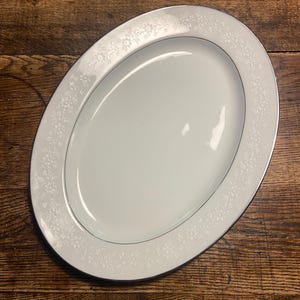 May include: Oval white serving platter with a floral design around the rim and a silver trim. The platter is on a wooden surface. The serving dish is a classic piece for serving food.