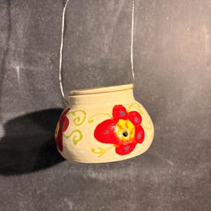 May include: A small, cream-colored ceramic pot with a lid and a thin silver wire handle. The exterior features hand-painted red flowers with yellow centers and green swirling vine accents. A small circular hole is visible on the front surface.