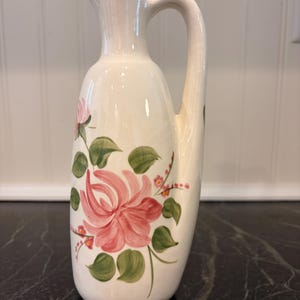 May include: A cream-colored ceramic pitcher featuring a hand-painted floral design with large pink flowers and green leaves. The pitcher has a tall, slender neck, a curved handle on the right side, and a small spout at the top for pouring liquids.