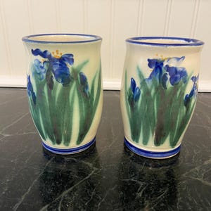 May include: Two ceramic tumblers with a cream-colored base, adorned with blue iris flowers and green leaves. The rims and bases are trimmed with blue. The tumblers are hand-painted and have a rustic, artisanal feel.
