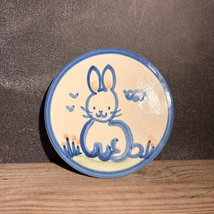 May include: A round ceramic plate with a cream-colored base and a blue rim. The center features a hand-painted blue line drawing of a seated rabbit with small orange accents on its ears and in the grass below. Two small blue clouds are visible.