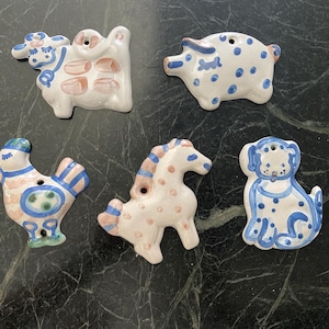 May include: A collection of five ceramic animal ornaments. The ornaments include a cow, pig, rooster, horse, and dog. Each ornament is hand-painted with a white base and blue and pink accents. Each has a hole for hanging.