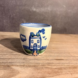 May include: A small, off-white ceramic cup featuring a hand-painted blue and green design of a house with a chimney emitting smoke, surrounded by a fence and clouds. The rim of the cup is accented with a thin blue line, and the base is rounded.