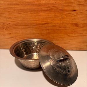May include: A round, hammered metal bowl with a matching lid. The bowl features a textured, dimpled surface throughout its interior and exterior. The lid has a smooth, polished finish with an engraved floral design and a small metal handle.