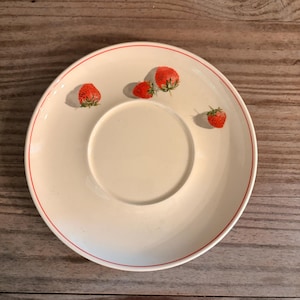 May include: A round, off-white ceramic saucer featuring a thin, circular red rim accent. Three small, fresh red strawberries with green stems are positioned on the upper portion of the saucer, casting soft shadows onto the smooth, glossy surface.