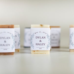 An assortment of mini hotel sized, wedding soap favors with white wraps.  The wraps have a floral edging design and the bride and groom's name is centered in the middle, with their wedding date above and the soap scent listed at the bottom.