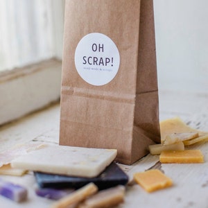 Könnte beinhalten: Eine braune Papiertüte mit einem weißen runden Aufkleber, auf dem "OH SCRAP! soap ends & scraps" steht. Die Tüte ist gefüllt mit verschiedenen kleinen Seifenstücken in verschiedenen Farben und Formen.