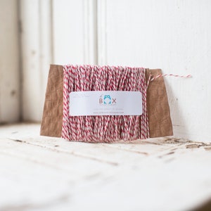 May include: Red and white baker's twine on a brown paper card. The card has a logo that says "the box", "craft supply", and "love the moments of giving" with the website "theboxboutique.com".
