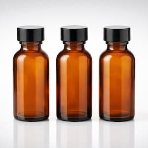 Op de afbeelding: Drie amberkleurige glazen flessen met zwarte doppen worden getoond tegen een witte achtergrond. De flessen zijn cilindrisch met afgeronde schouders en een iets bredere basis. De doppen zijn plat en cirkelvormig. De flessen zijn leeg en reflecteren het licht.