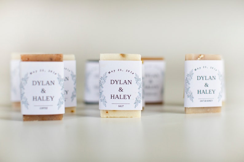 An assortment of mini hotel sized, wedding soap favors with white wraps.  The wraps have a floral edging design and the bride and groom's name is centered in the middle, with their wedding date above and the soap scent listed at the bottom.
