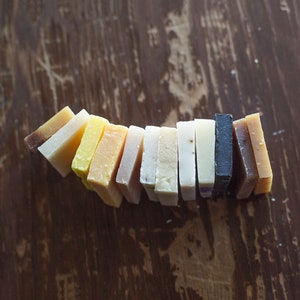 May include: A selection of handmade soap bars in a variety of colours and textures, arranged in a curved line. The soaps range from light yellow to dark brown, with visible natural ingredients. The background is a rustic, dark wooden surface.
