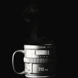 May include: A black and silver camera lens with a white label that reads "AUTO FOCUS 310ml". The lens has a silver ring with numbers and markings. The lens is sitting on a dark surface with steam rising from it.