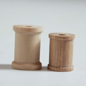 May include: Two wooden spools, one larger than the other, both with a smooth, light brown finish. The spools are made of wood and have a hole in the center.