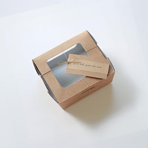 May include: A square, brown cardboard bakery box with a clear window on top. The box is tied with twine and has a small tag that reads "pure lovin' from the oven."