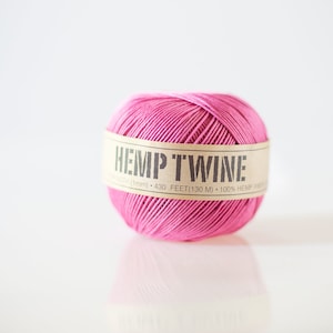 May include: A ball of pink hemp twine with a tan label wrapped around it. The label reads "HEMP TWINE" and includes the length of the twine, 130 metres. The twine is made of 100% hemp fibre.