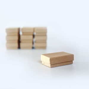 May include: A small, brown cardboard box sits on a white surface. A stack of similar boxes is blurred in the background.