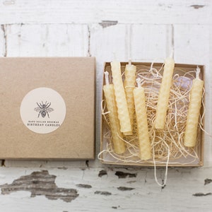 May include: A set of hand-rolled beeswax birthday candles in a kraft paper box. The box features a round label with a bee illustration and the text "Hand Rolled Beeswax Birthday Candles". The candles are a light yellow color.