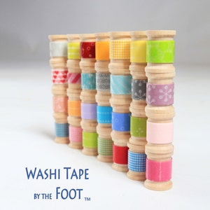 May include: A collection of colourful washi tapes wound around wooden spools. The tapes feature various patterns, including hearts, snowflakes, and geometric designs. The text "WASHI TAPE BY THE FOOT™" is printed below the spools.