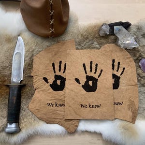 May include: An assortment of items on a fur-like surface. Includes a knife with a black handle, three pieces of tan paper with black handprints and the text "We know", a brown leather pouch, and crystals.