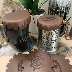 May include: Two drinking mugs with brown leather lids and leather straps. One mug is black and horn-shaped, the other is silver with text from the First Amendment. Brown leather coasters and a bottle opener are also visible.