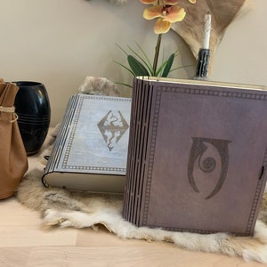 May include: Two wooden book-shaped boxes with engraved designs. The box on the left has a white background with a dragon symbol and the text "Skyrim". The box on the right has a brown background with a symbol of a dragon with a circle in its eye.