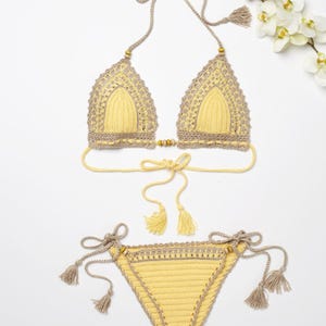 May include: A yellow crochet bikini with brown trim and tassels. The bikini top has a triangle shape and ties at the neck and back. The bikini bottom has a low-rise cut and ties on the sides.