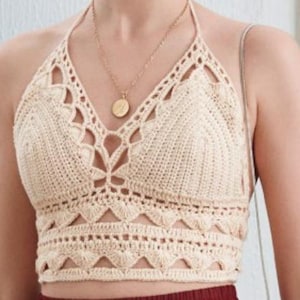 May include: Cream-colored, handmade crochet halter top. The top features a V-neck design with intricate openwork patterns and a scalloped hem. A gold-colored necklace with a pendant is visible. The top is a stylish piece of clothing.