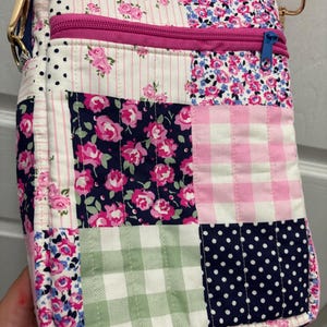 Puede incluir: Un bolso bandolera patchwork con una variedad de estampados florales, cuadros y lunares en rosa, blanco, azul marino y verde. El bolso tiene una cremallera rosa, una correa rosa y herrajes dorados.