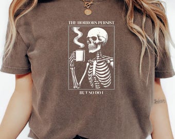 Może przedstawiać: Brązowa koszulka z grafiką przedstawiającą szkielet trzymający kubek kawy. Pod grafiką znajduje się napis "The horrors persist but so do I".