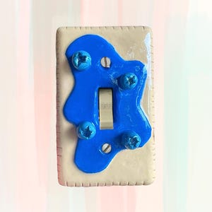 May include: A custom light switch plate with a blue abstract design. The plate is off-white with a blue, organic shape overlay. Four blue screws secure the blue shape. The switch is in the "OFF" position.