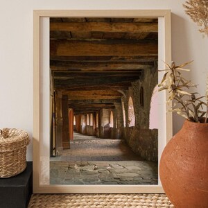 May include: Framed print of a stone corridor with a wooden ceiling. The corridor has arched windows and doors, with sunlight streaming through. The print is in a light wood frame, displayed on a woven rug with a basket and vase.