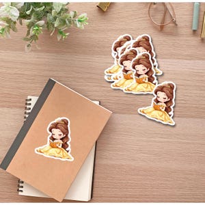 Puede incluir: Pegatinas que representan a una princesa de dibujos animados con un vestido amarillo leyendo un libro. Varias pegatinas est&aacute;n dispuestas sobre una superficie de madera, junto a un cuaderno y vegetaci&oacute;n.
