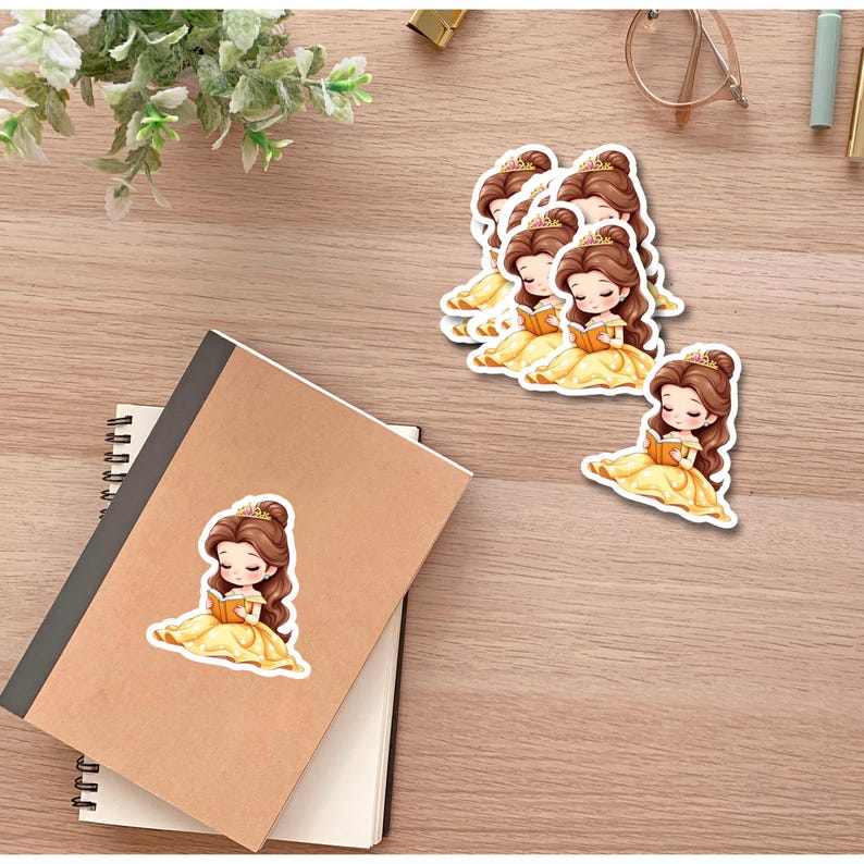 Puede incluir: Pegatinas que representan a una princesa de dibujos animados con un vestido amarillo leyendo un libro. Varias pegatinas est&aacute;n dispuestas sobre una superficie de madera, junto a un cuaderno y vegetaci&oacute;n.